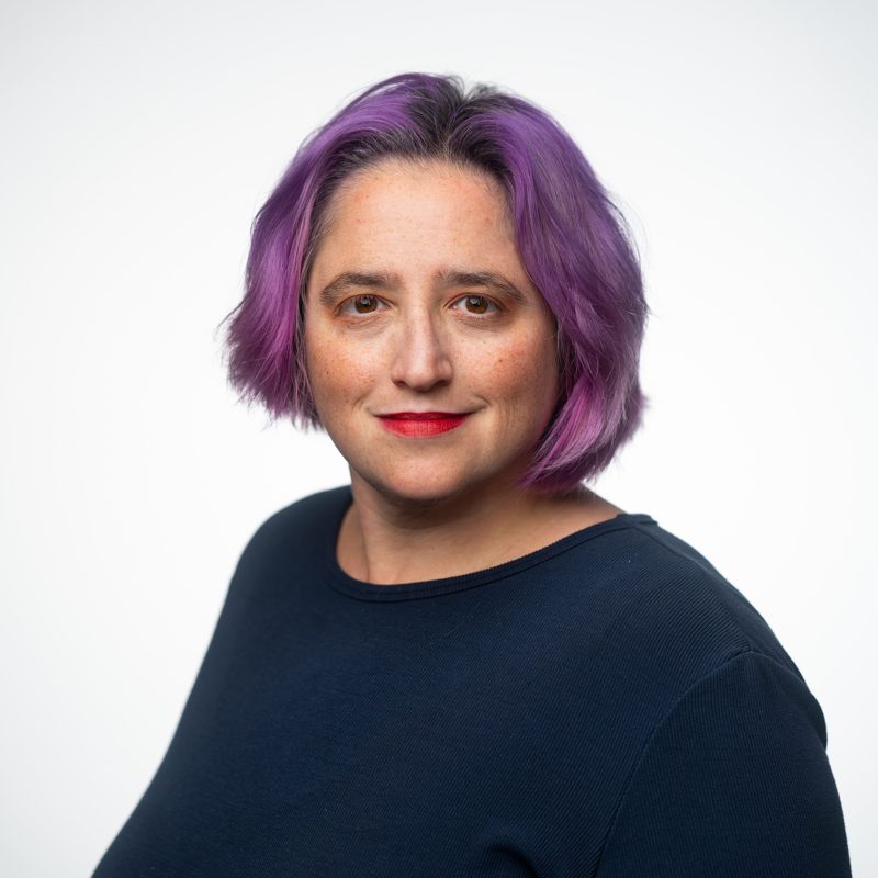 Michelle Brenner, smiling, with chin-length purple hair, red lipstick, and a navy top.